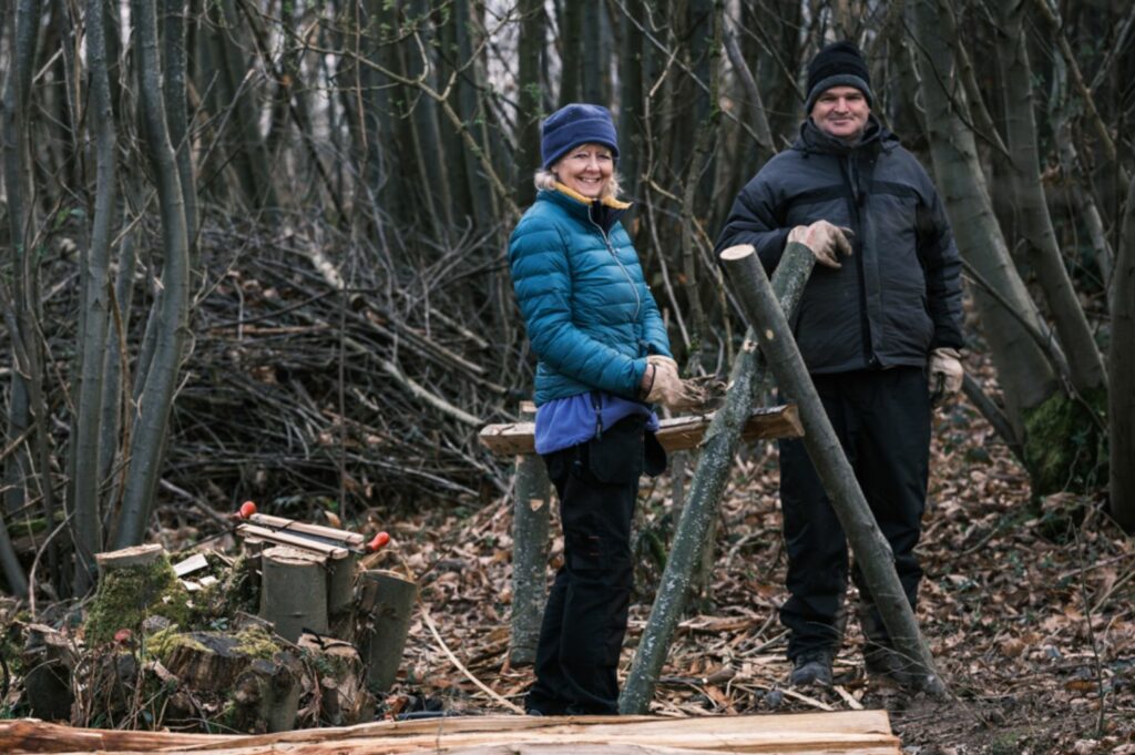 Two people smiling and standing in the woods with logs around them.