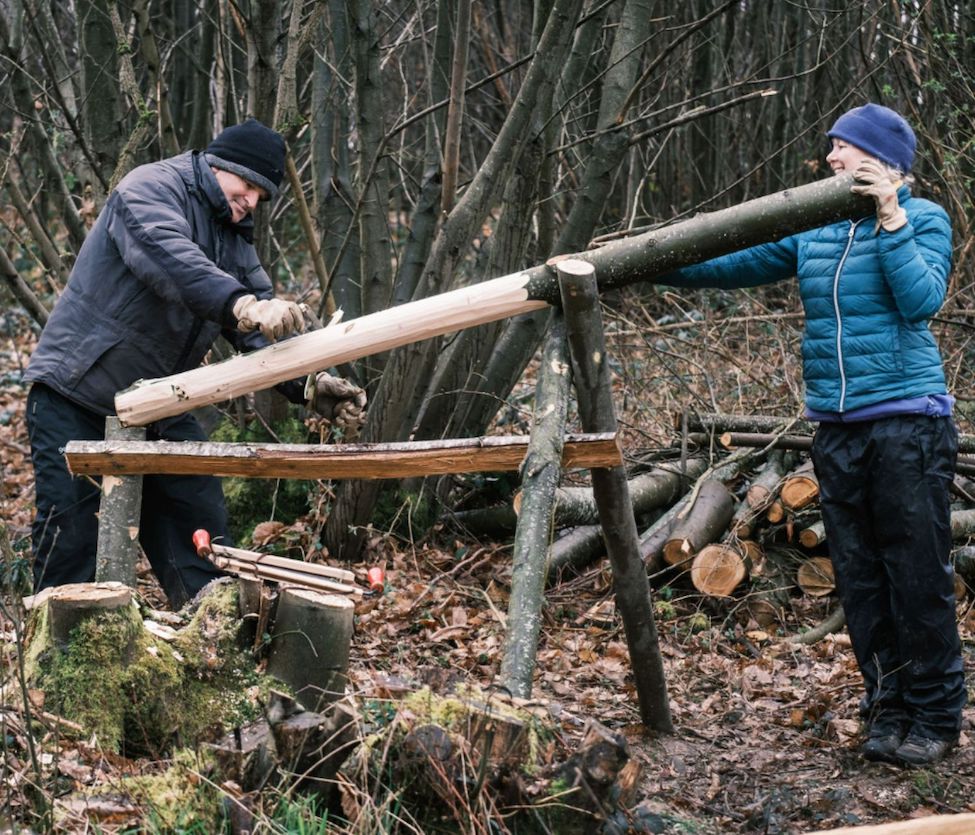 Two person working on a log, one is shaving the wood log and the other holding the log in the woodland.