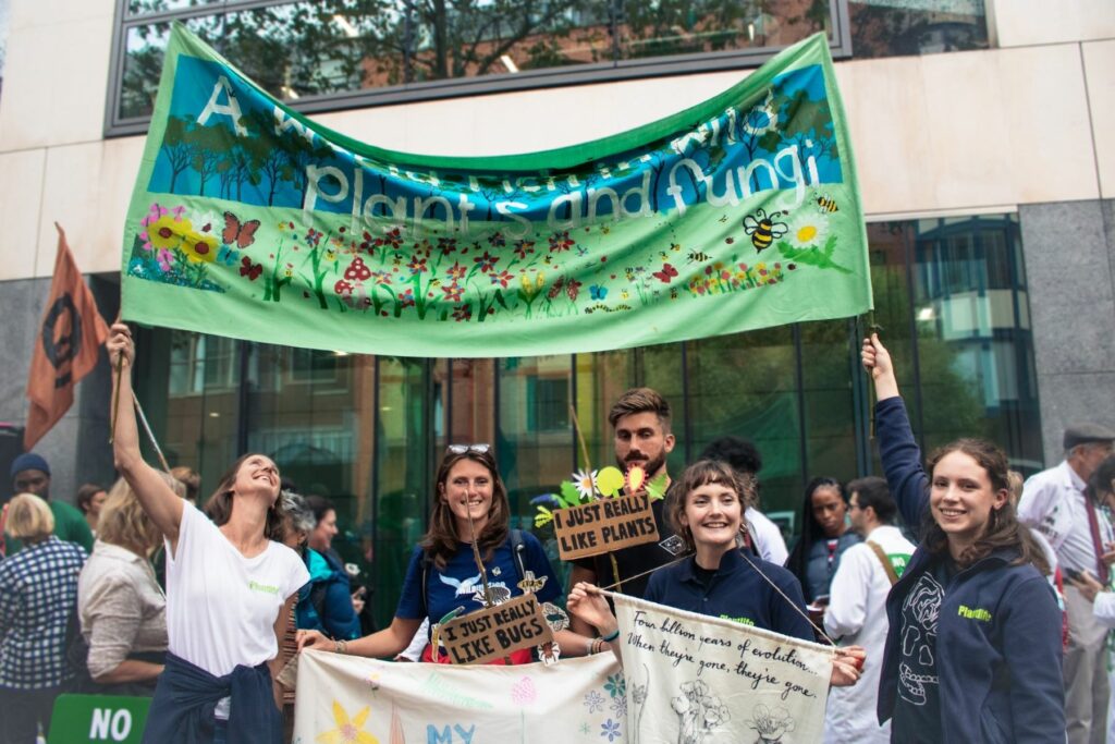 A team of people wearing Plantlife uniforms under a banner which reads 'A world rich in wild plants and fungi'
