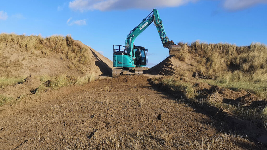 A blue digger is carving a path between two grassy dunes