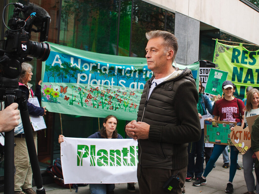 Chris Packham stood in front of a professional camera crew with handmade Plantlife protest banners carried by staff at a protest