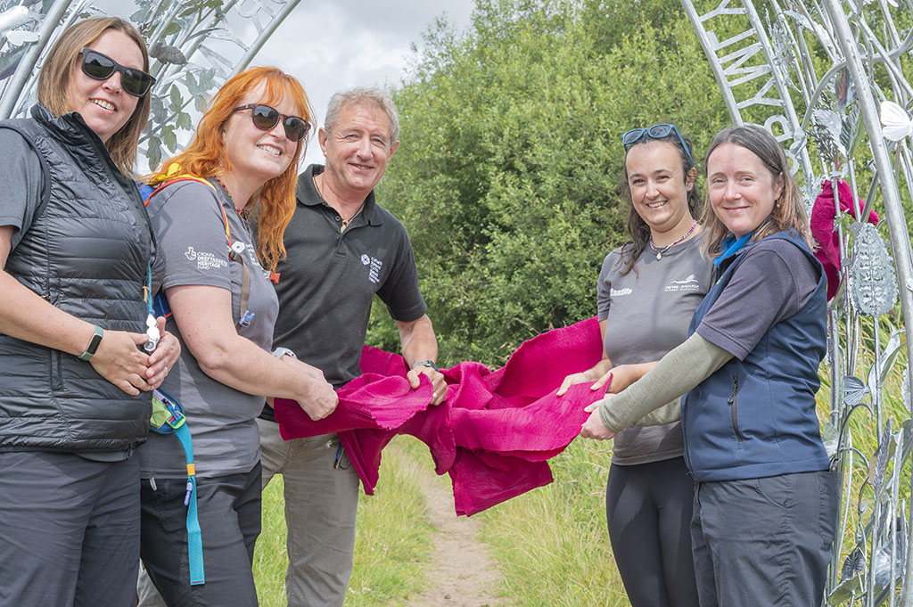 Four women and one man smiling holding a burgundy ribbon that is tied around a metal sculpture