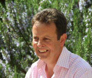 Headshot of David Hill in a pink checked shirt in front of a green bush