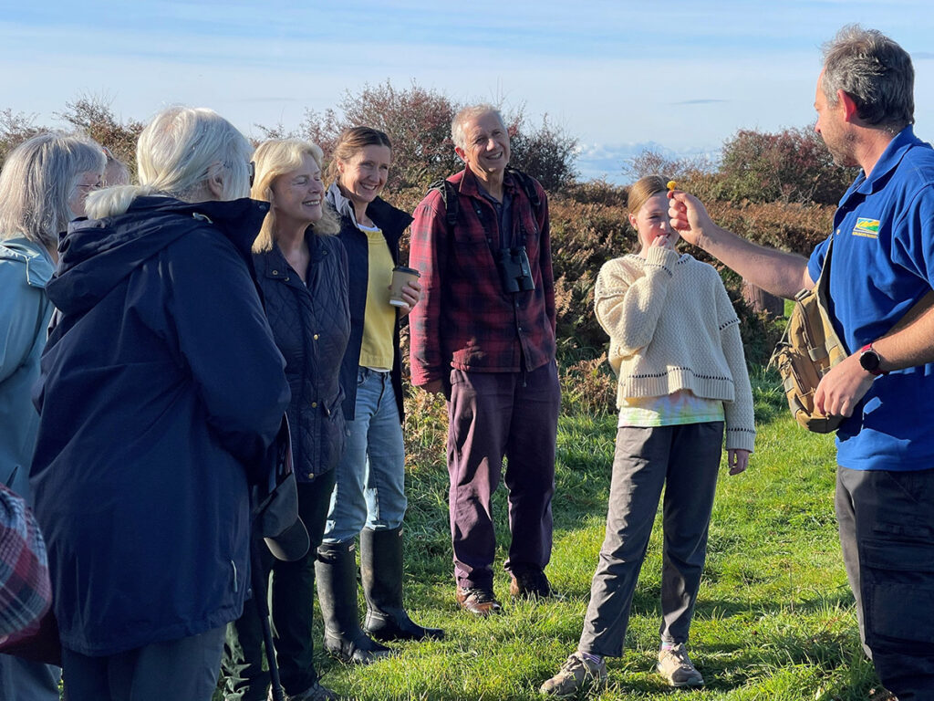 A group of people stood in a semi circle in a field watching and listening to a man in a blue tshirt. The man is holding a mushroom.