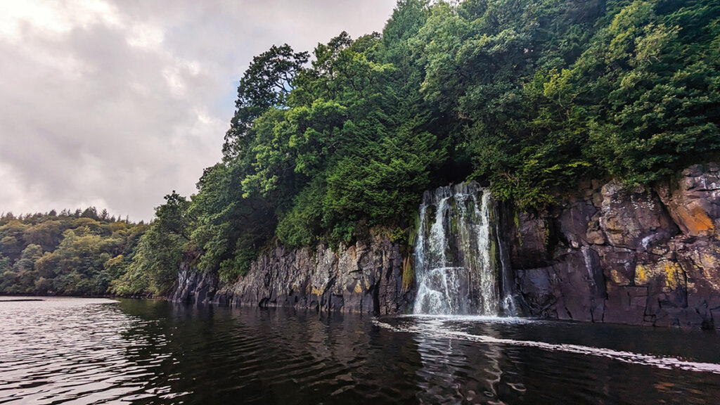 A waterfall falling across a rockface with green trees above the rocks and a large body of water below