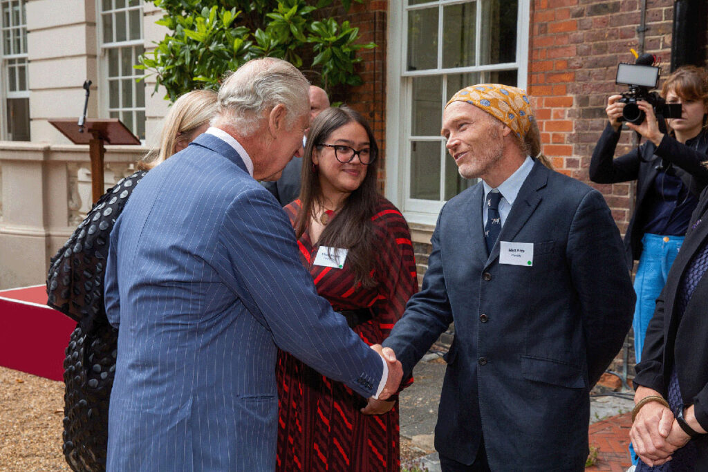 Matt Pitts wearing a suit and an orange head scarf shakes hands with King Charles