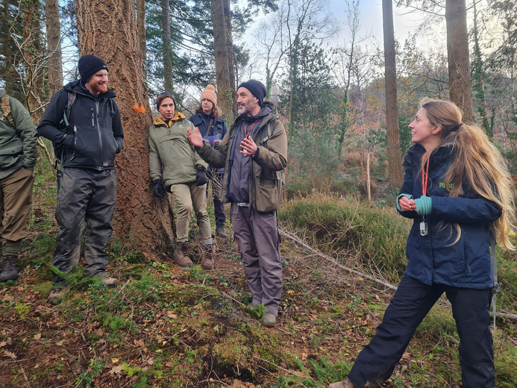 A group of people wearing winter clothing stand listening to a man talk inside a forest