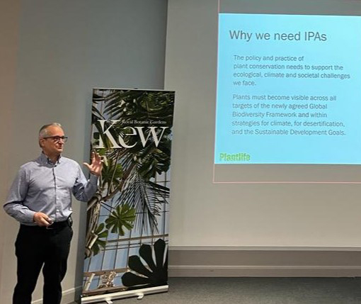 Ian Dunn delivering a presentation wearing professional clothes stood in front of a Kew roller banner and a PowerPoint is being projected onto the wall.