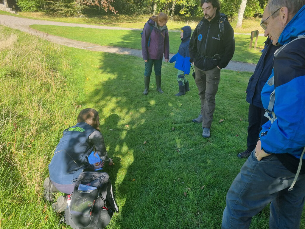 A person in Planlife uniform is kneeling on grass while a group of people stand around looking and listening