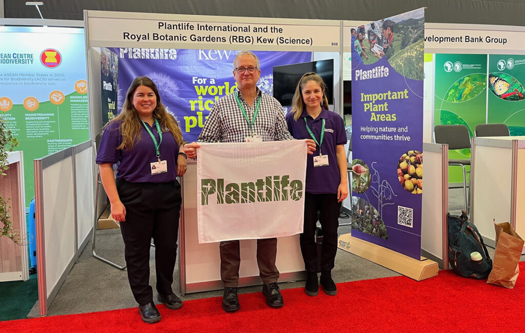 Ian Dunn, Claire Rumsey and Jo Riggall pose in front of a Plantlife event booth with a smile and holding a white Plantlife flag