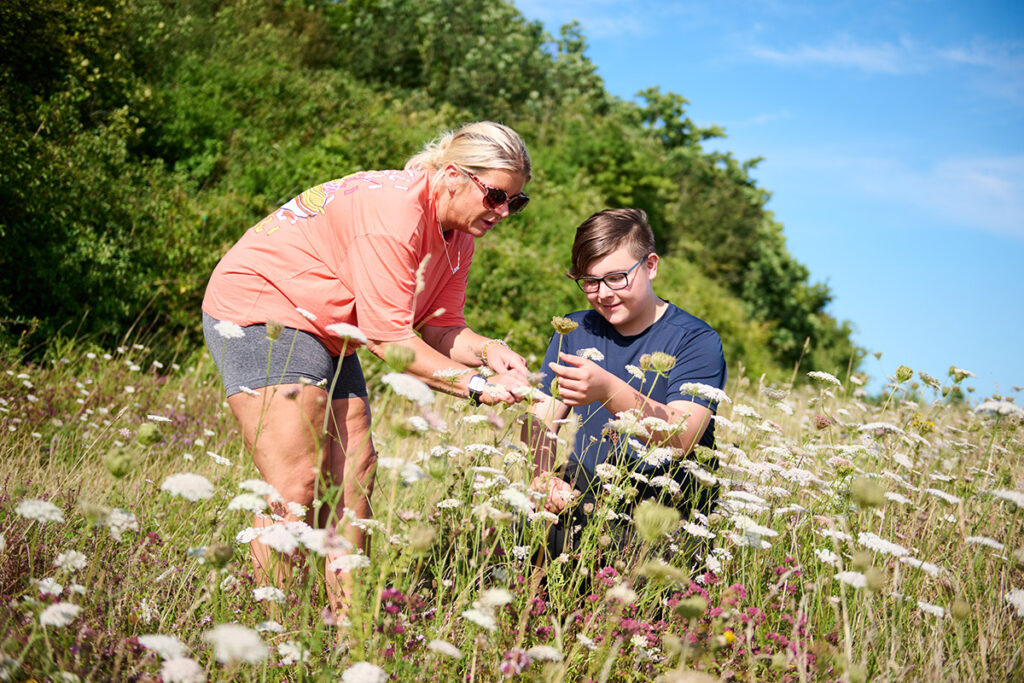 A teenager and a woman looking closely at a wildflower whilst crouching low in a wildflower field on a sunny day