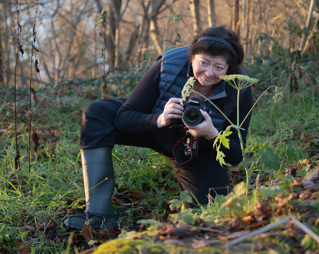 Sue McBean crouching low to the ground with a Canon camera in hand, pointing it towards a flower