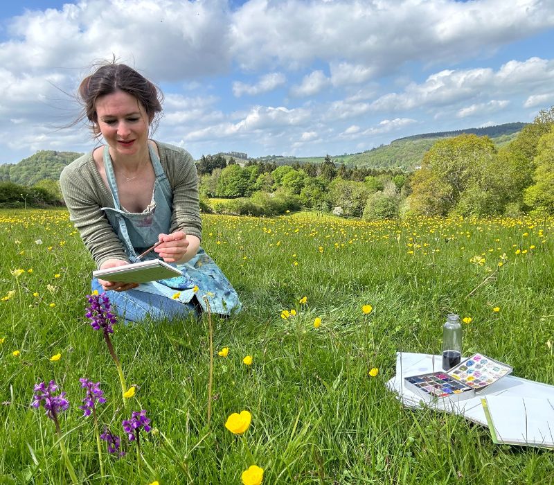 a person in a meadow looking at yellow and purple flowers with painting materials