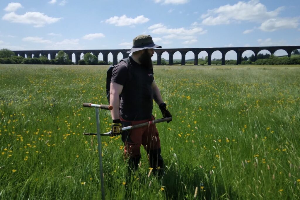 A man holds tools in the middle of Seaton Meadow, ready to start meadow management work