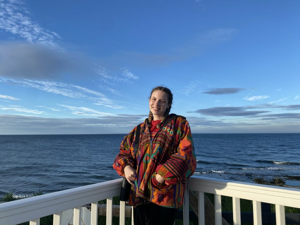 Plantlife staff member Caitlin Thomas stands facing the camera. She is next to a white fence with the sea behind her on a clear day.