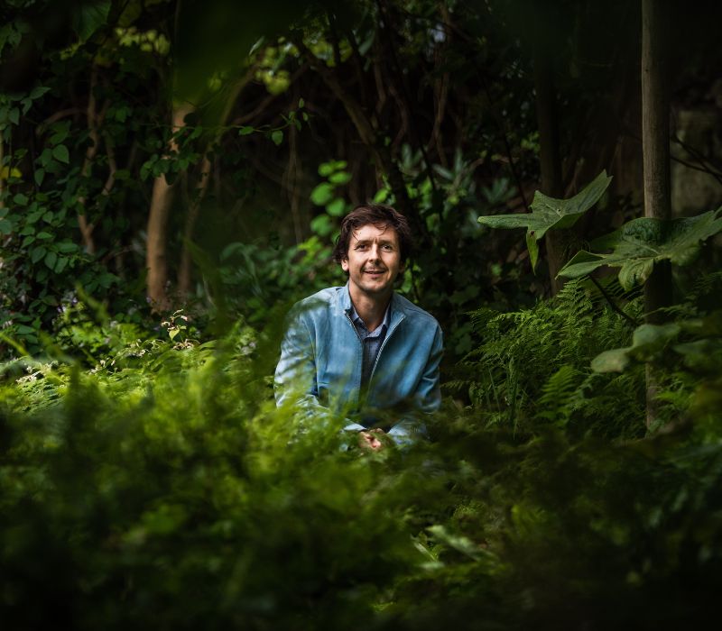 Landscape Designer and creative Gardener, Dan Bristow, sits in the centre of a dark garden, surrounded by rich green plants included beautiful leafy ferns