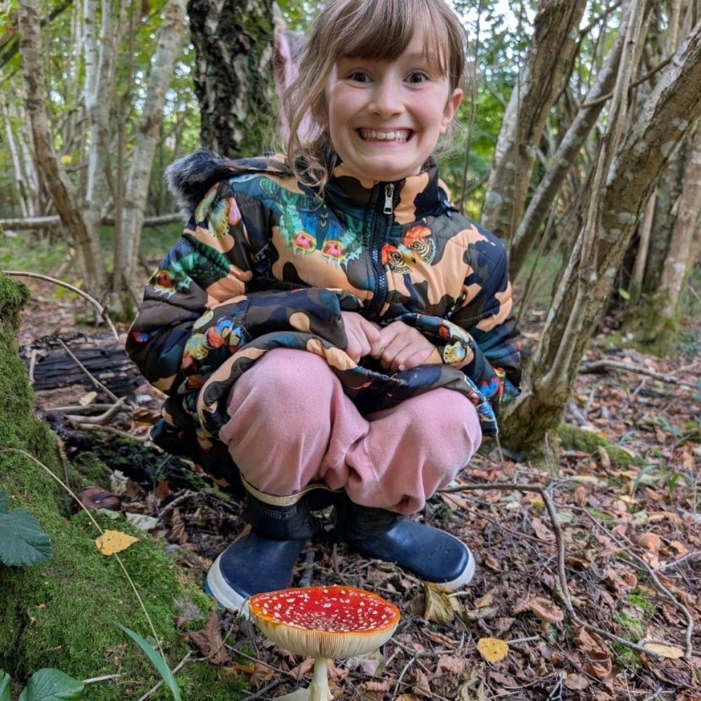 A young girl kneels on the forest floor next to a Fly Agaric fungus