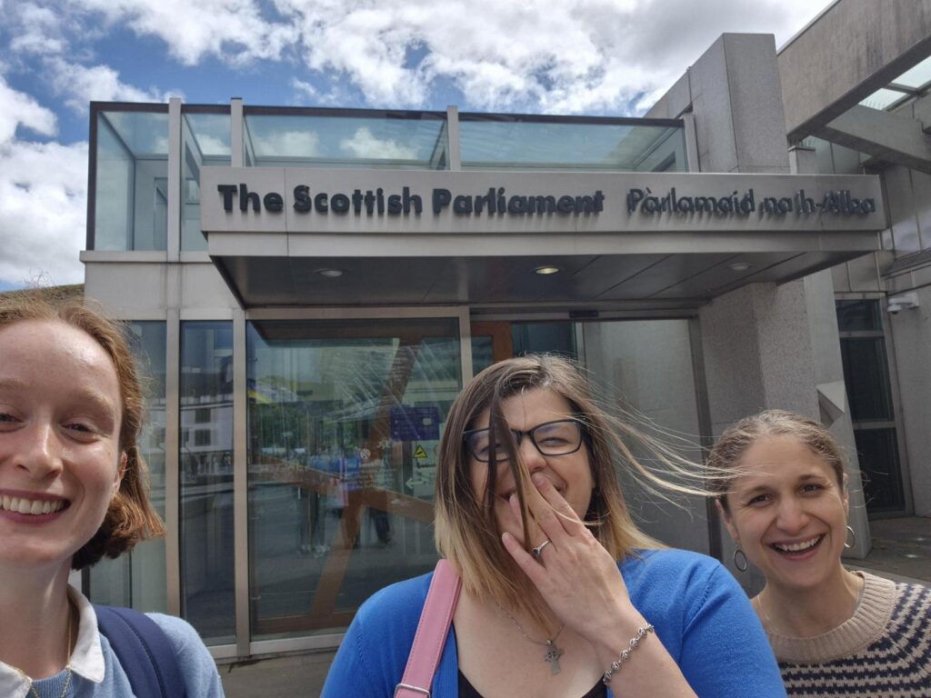 3 Women smile standing outside The Scottish Parliament building