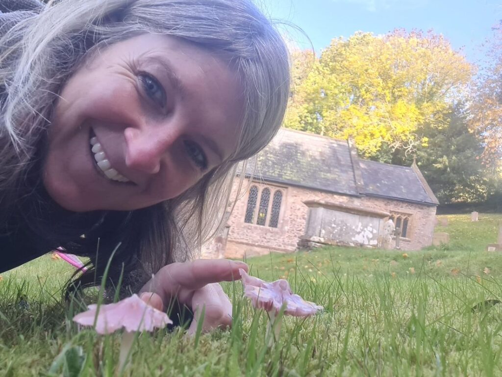 A woman leans close to the ground to point out a pretty Pink Waxcap mushroom