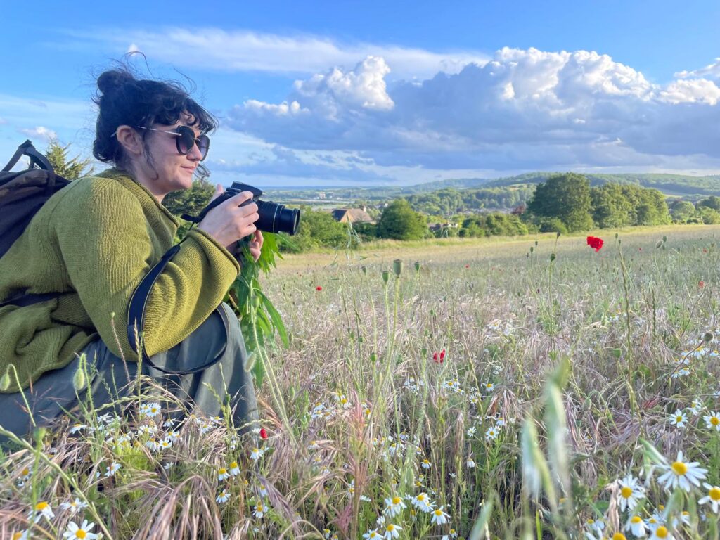 A woman, Ayesha, with sunglasses on and wearing a green jumper and grey trousers. She has black hair tied up with a fringe and is crouching in a field of poppies at Ranscombe, holding a camera.