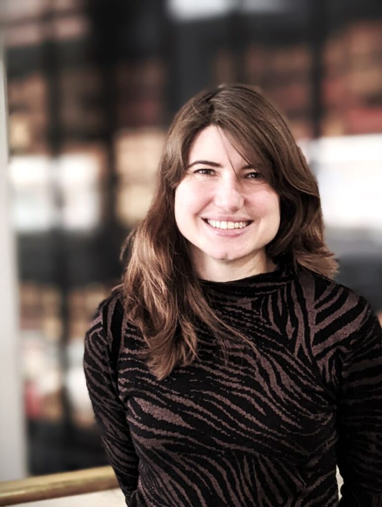 Photograph shows a professional headshot of a woman smiling at the camera. She has mid length brown hair and is wearing an abstract striped top in brown and black.