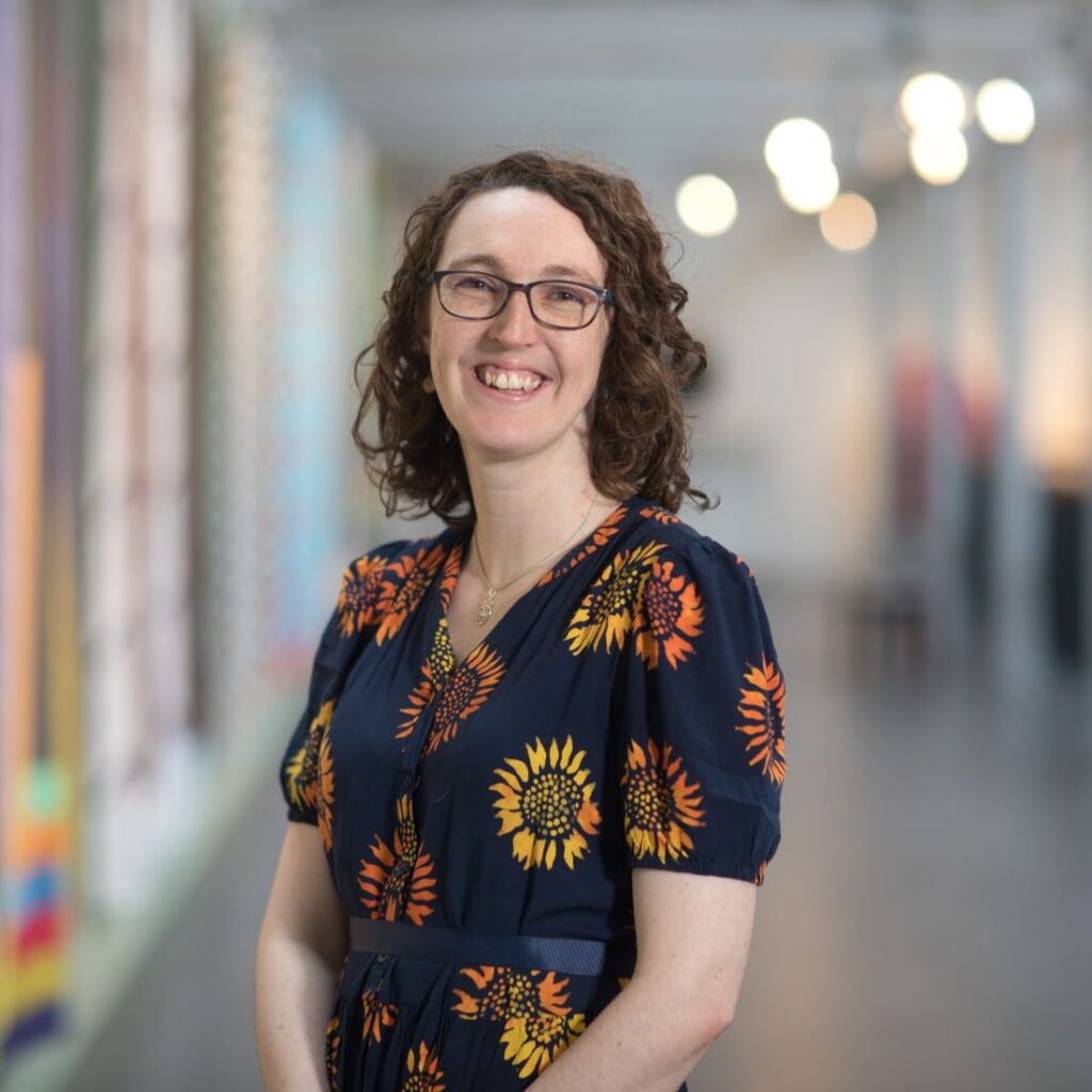Photograph shows a professional headshot of a woman. She has brown shoulder length hair, black rimmed glasses and is wearing a dark blue top with yellow flowers on it. She is smiling.