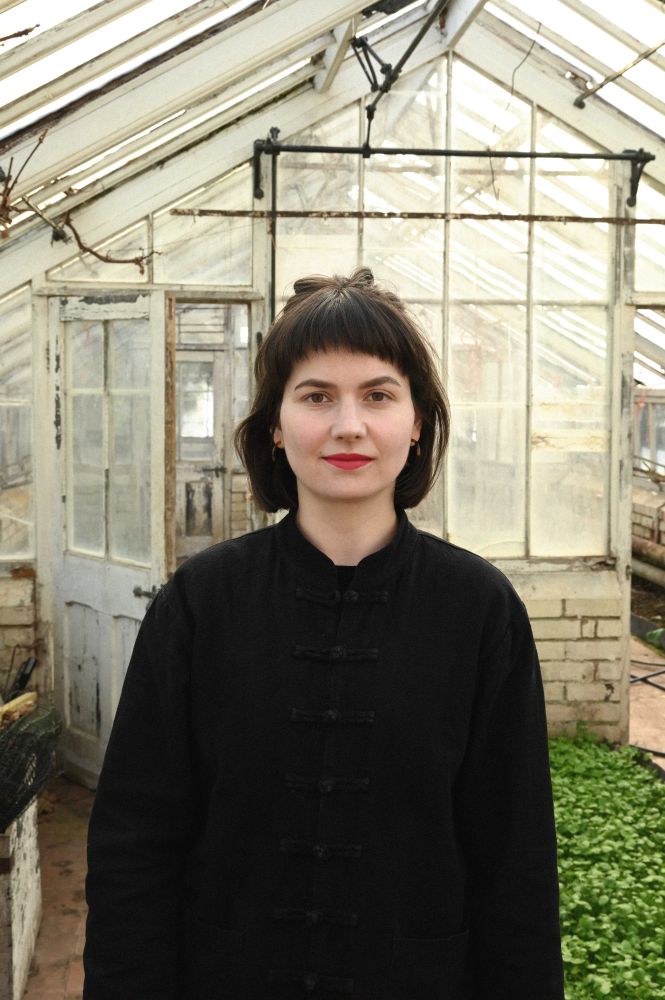 Photograph is a headshot of Caroline Vitzthum. She has dark short hair with a fringe, and wears a black long sleeve shirt. She is standing in a greenhouse.