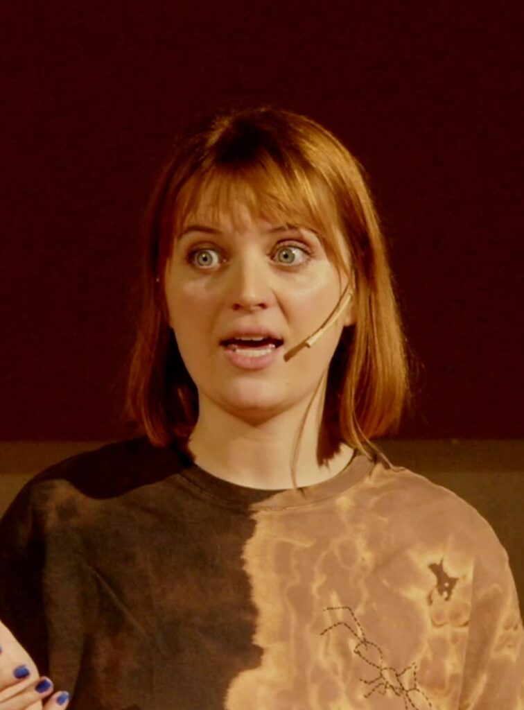 Photograph is a headshot of Frankie Tuck. She has light brown hair in a bob style and is wearing a brown t shirt with a tie-dye pattern.