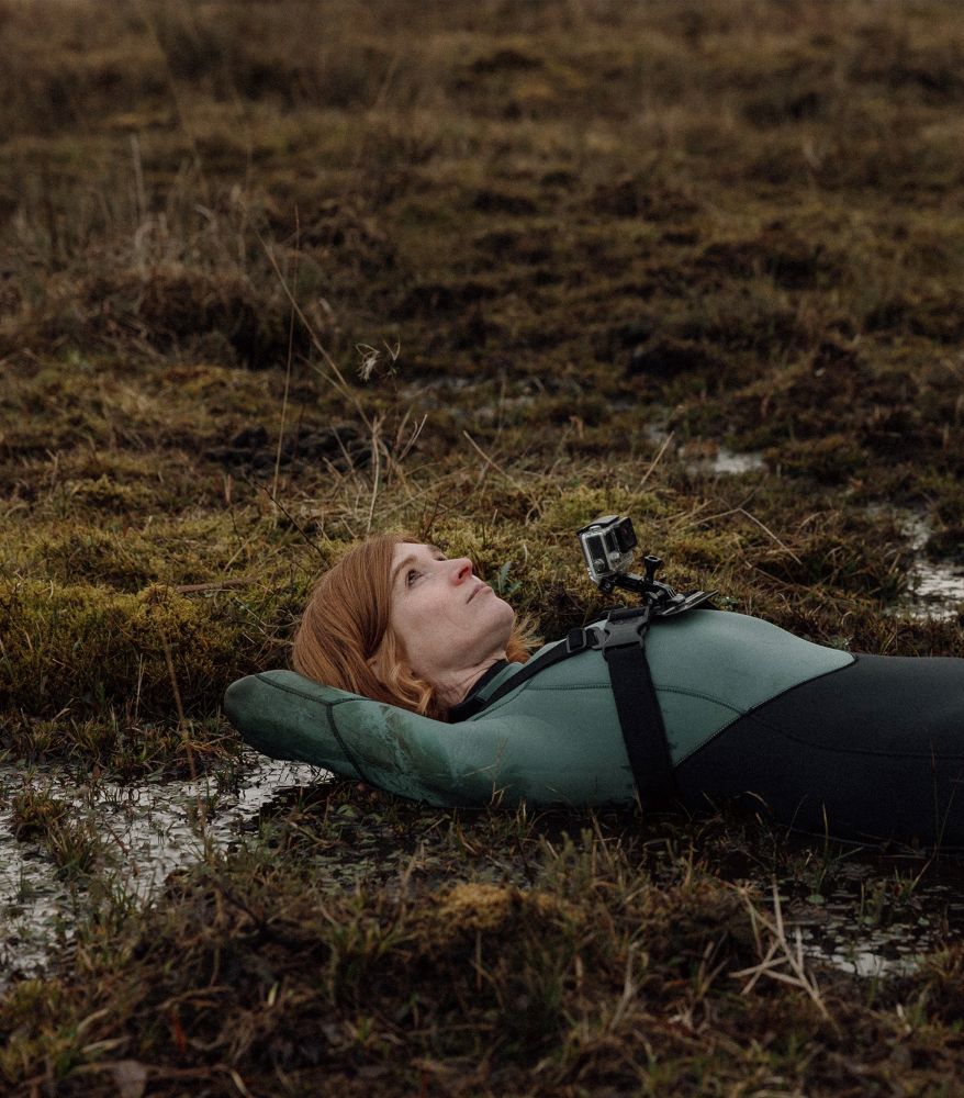 Photograph shows a woman lying down on her back in a peatland, looking up at the sky