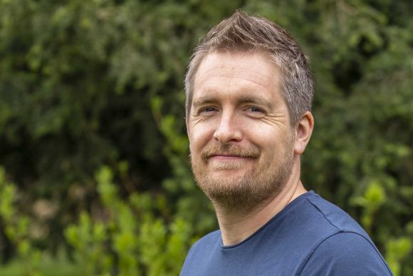 Photograph is a headshot of a man standing in front of trees that are blurred in the background. He wears a blue t shirt.