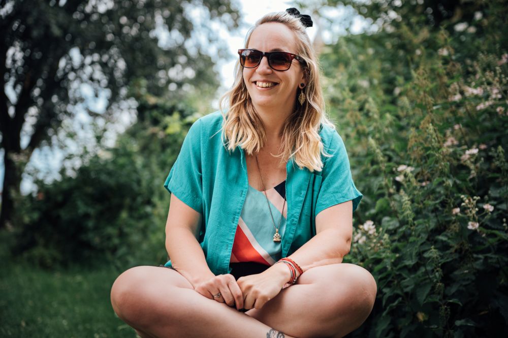 Photograph shows a woman sat with her legs crossed in an outdoor setting. There are blurred trees in the background. The woman wears a bright blue short sleeve shirt, shorts and a sunglasses.