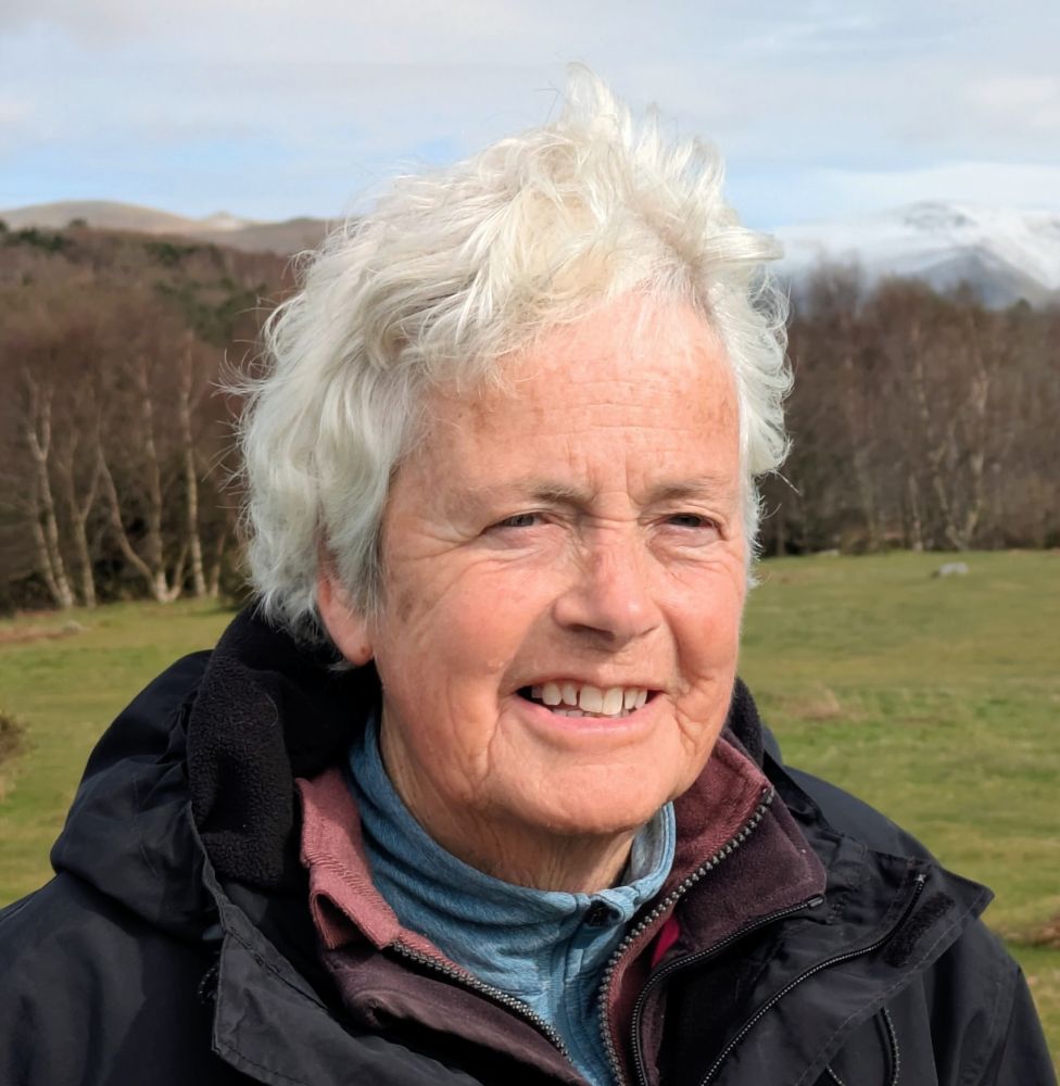 Photograph shows a close up of a woman standing in a grassland with trees in the background