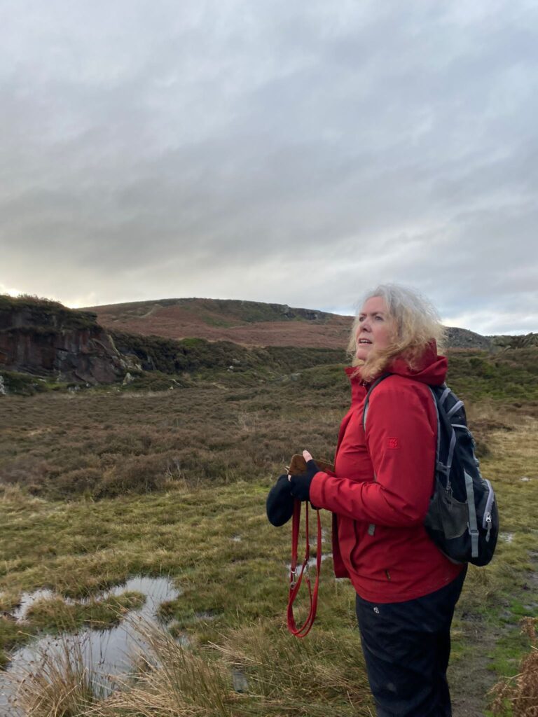 A woman stood in a moorland-type landscape wearing a red coat.