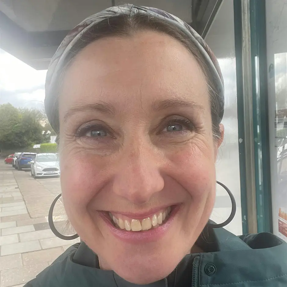 Photograph shows a close up of a womans face smiling. She is wearing hoop earrings, a pale green patterned head band and a green scarf.