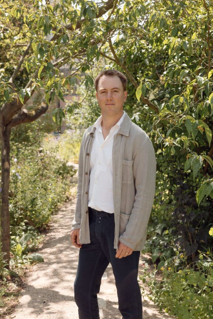 Photograph shows a man standing on a path surrounded by greenery, trees and plants. The man is wearing black trousers, a white collared top and a beige cardigan.