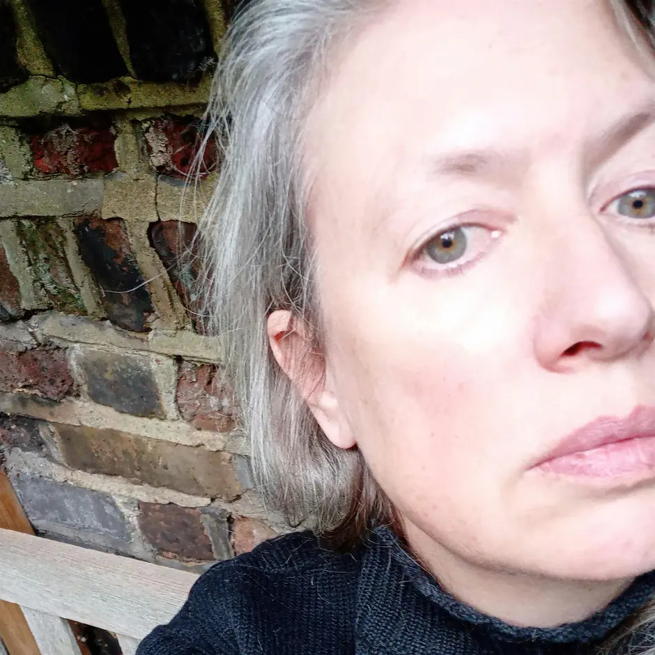 Photograph shows a close up of a woman's face, she has grey hair, blue/grey eyes and is wearing a black top