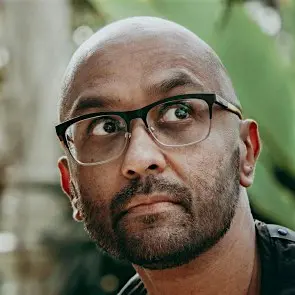 Photograph shows a close up of a man's face, he is wearing glasses and a black top