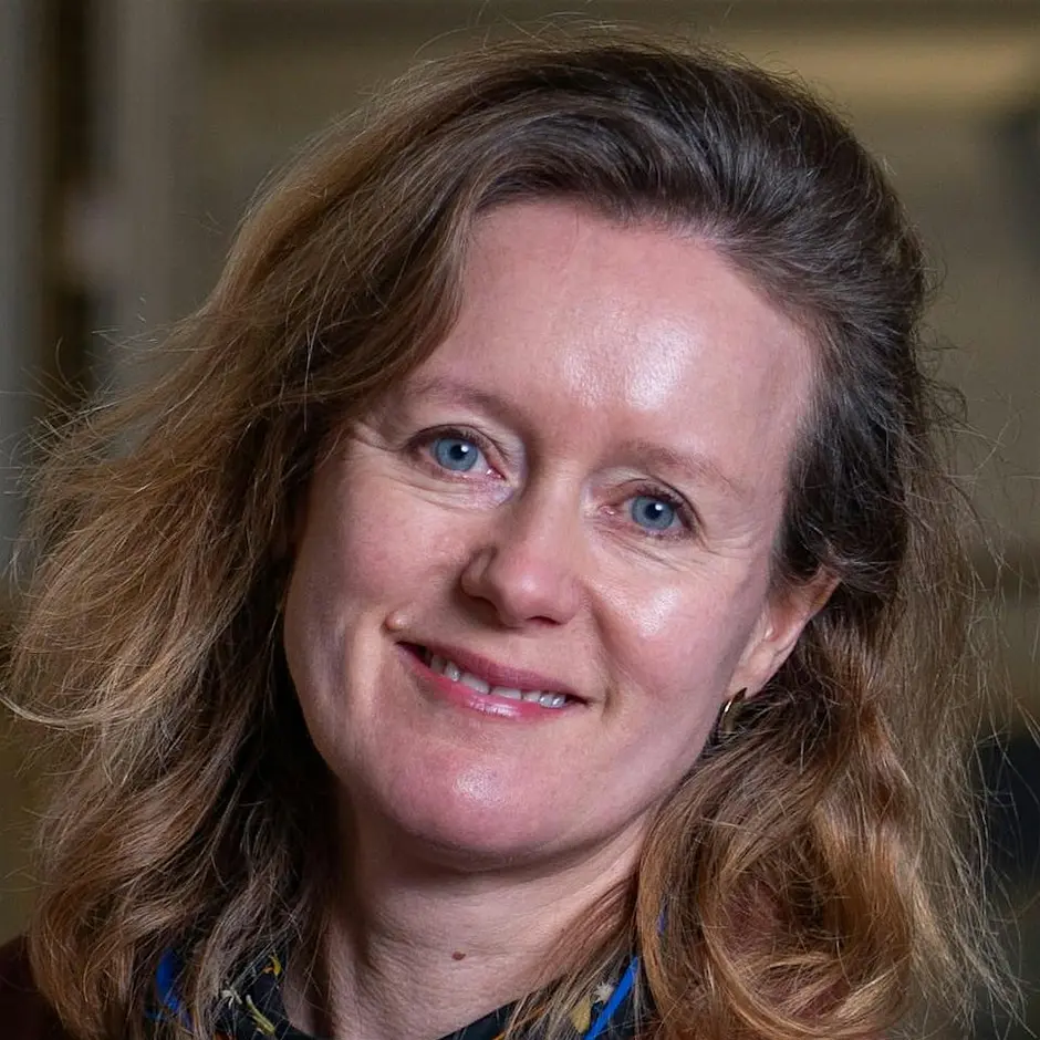 Photograph shows a close up of a woman smiling. She has long dark blonde hair and blue eyes.