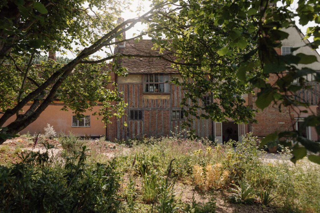 Photograph shows the house and garden of garden of artist and plantsman Cedric Morris. The house is partially visible through trees, and many plants are seen in front of it.