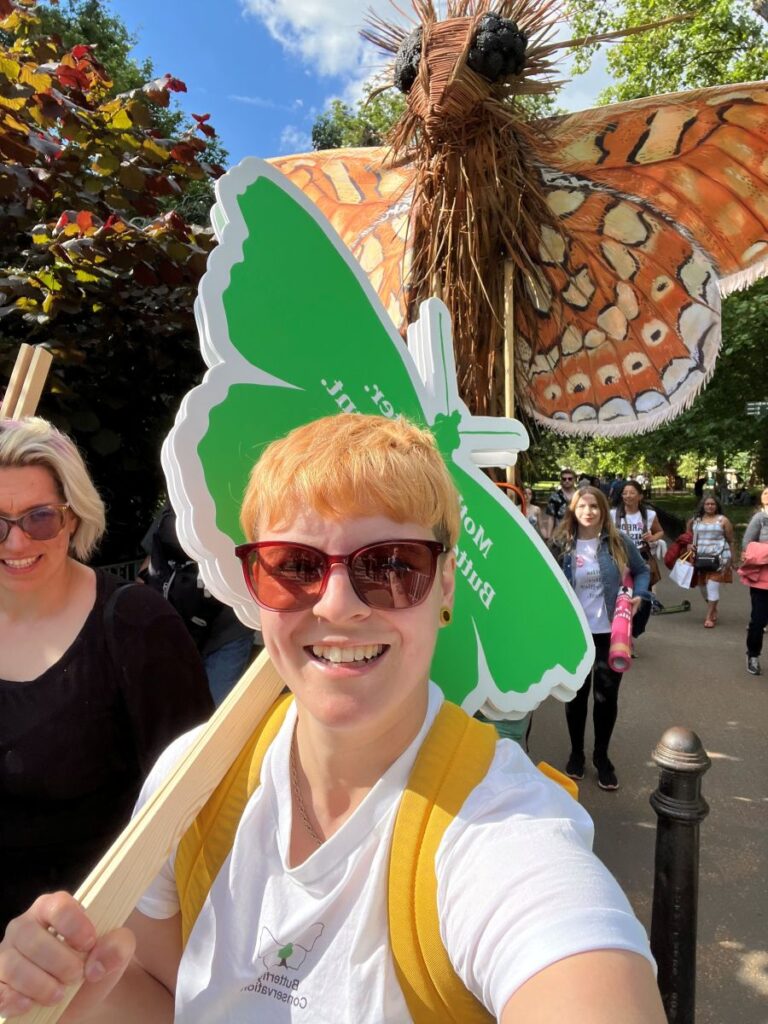 Photograph shows a woman holding a plaque during the Restore Nature Now March in London. The woman is wear a white top, yellow backpack and sunglasses.