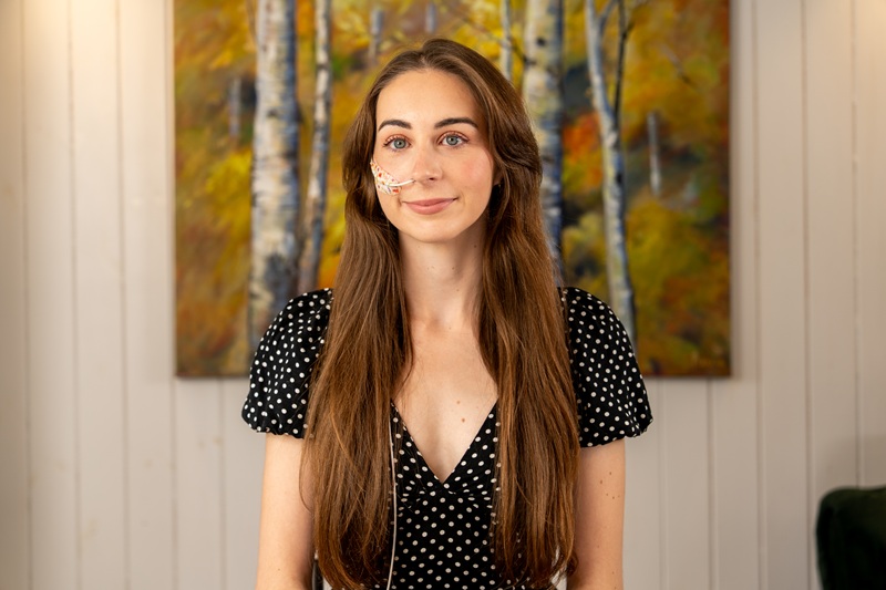 Photograph shows a woman standing in front of a painting on a white wall. The woman has long brown hair and is wearing a black top.